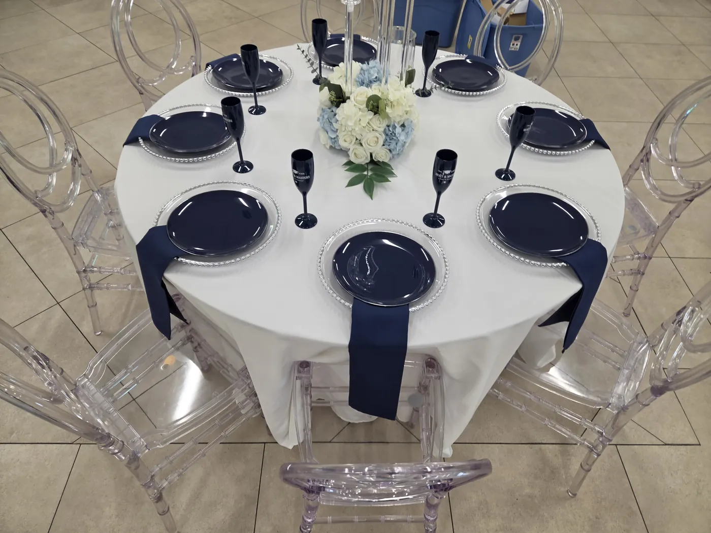 Round table set with white linen, navy chargers and napkins, glassware and ghost chairs